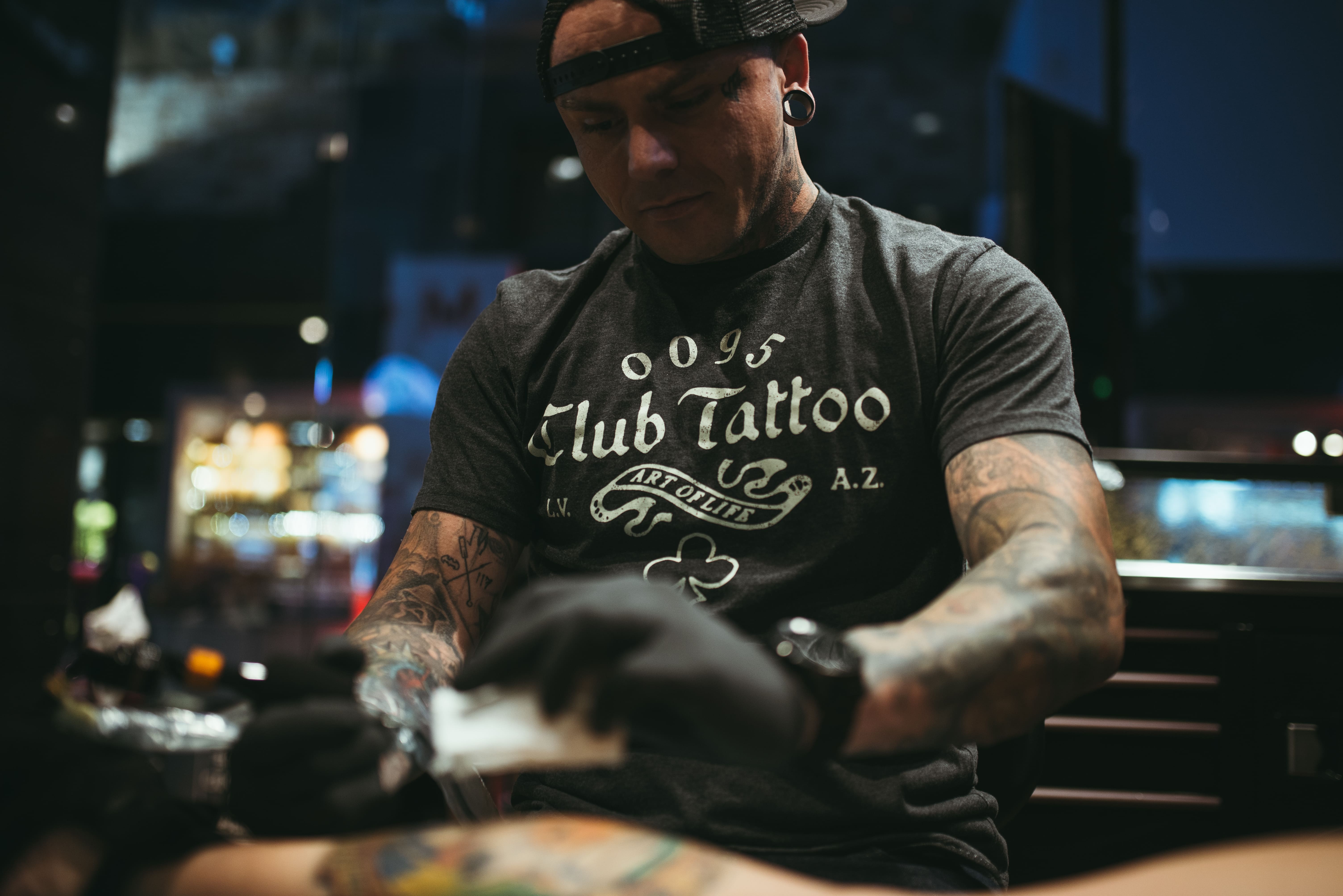 A close-up shot of a professional Club Tattoo artist wearing a black branded graphic t-shirt and gloves, meticulously working on a colorful custom tattoo in a clean, high-end studio setting.
