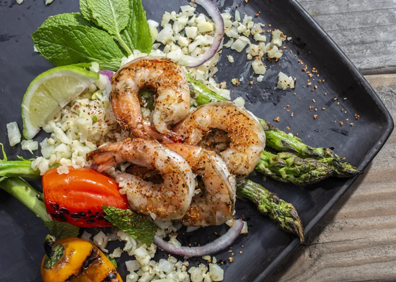 Grilled shrimp with asparagus, mint, lime, and diced vegetables on a black plate