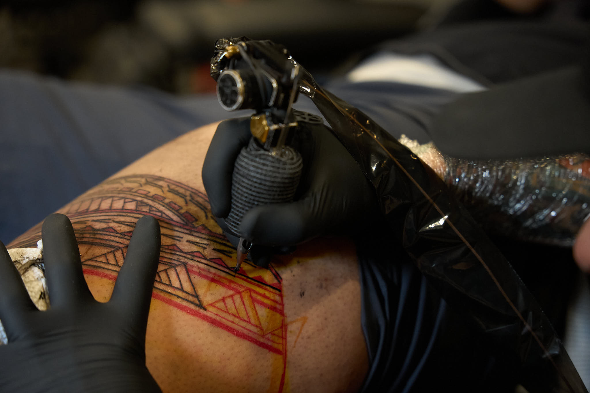 A close-up shot of a tattoo artist wearing black gloves using a tattoo machine to outline a geometric tribal pattern on a client's skin, with red stencil guidelines visible underneath.