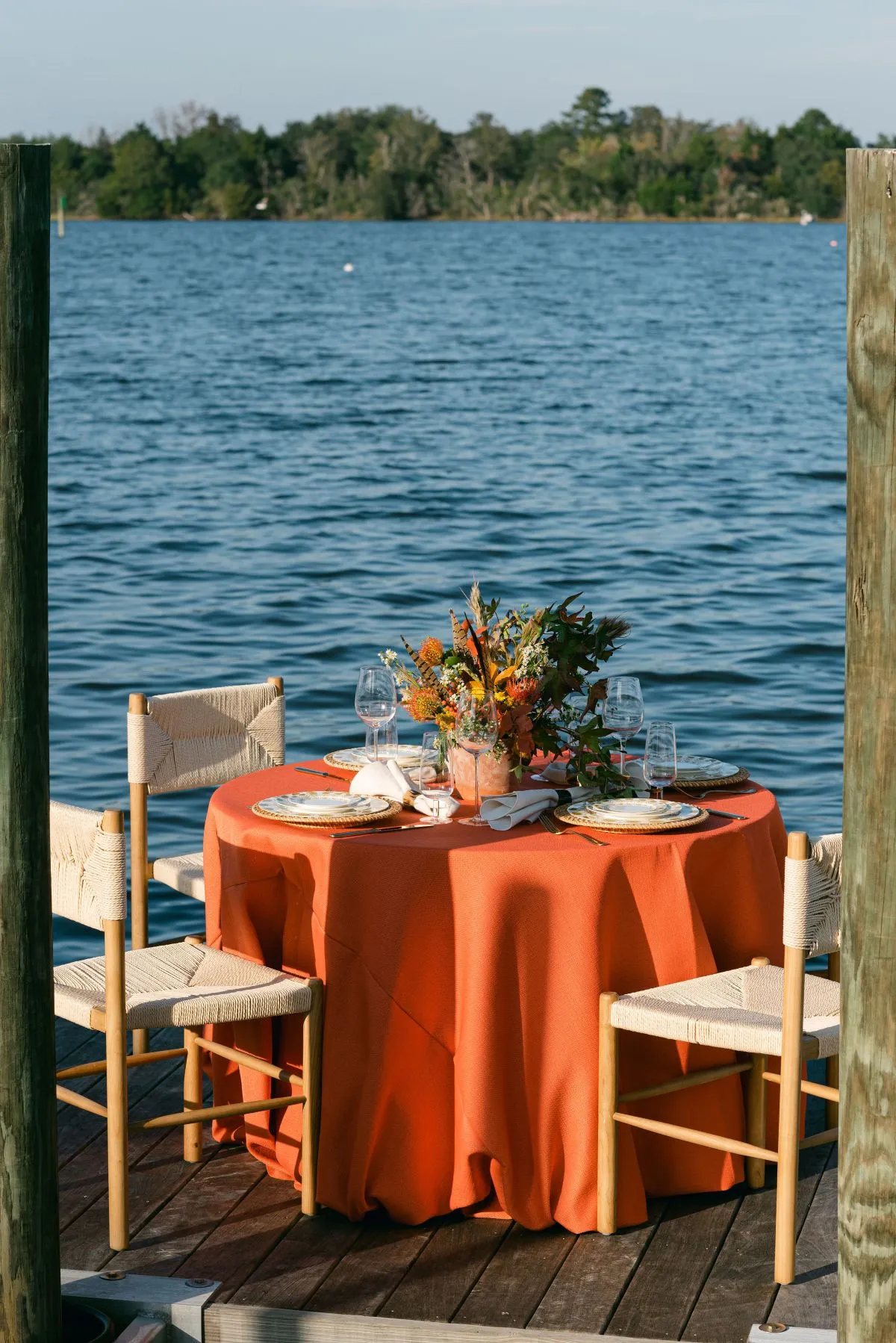 Round waterfront dining table with rust-orange linen, woven chairs, and floral centerpiece.