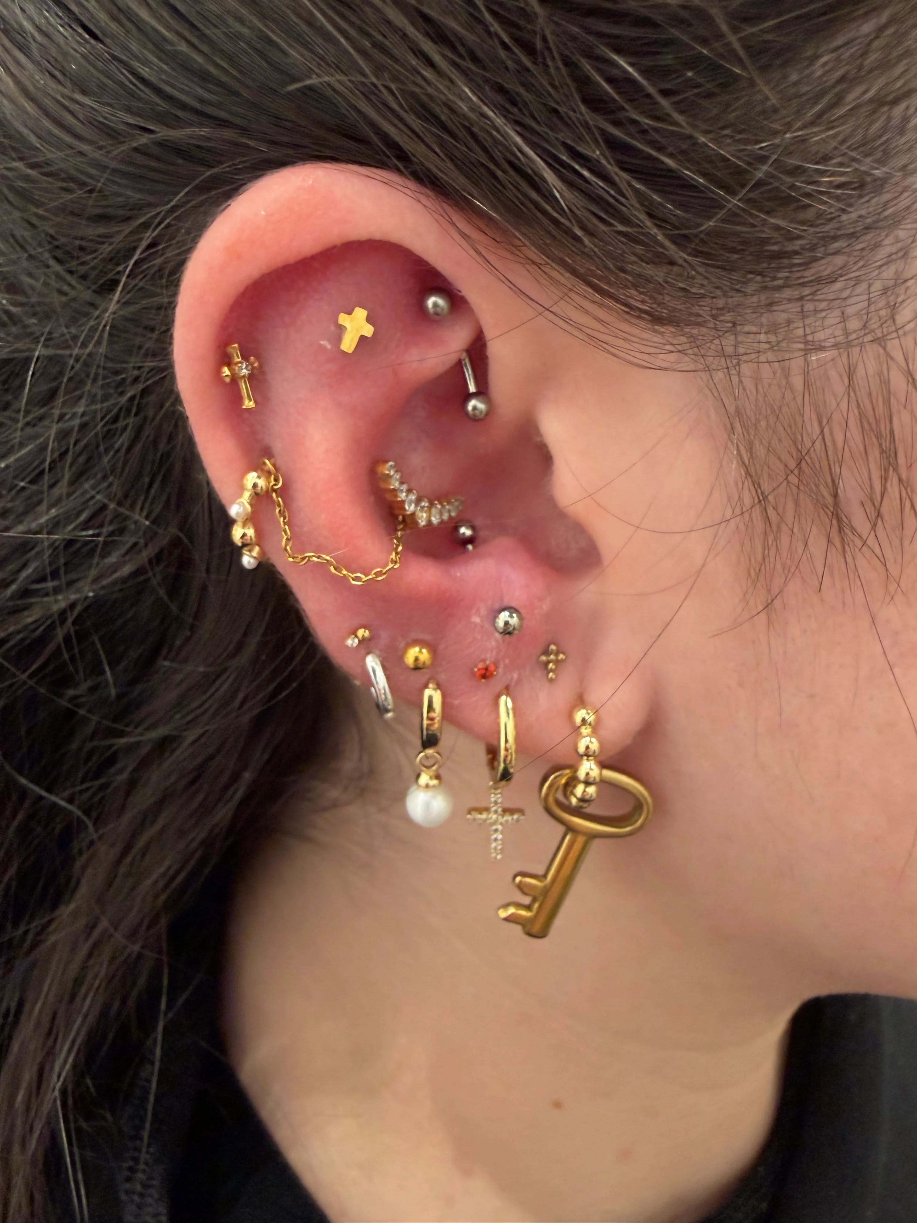 Close-up of an ear showcasing a maximalist gold jewelry collection. A vertical barbell sits in the rook, a gold chain drapes across the mid-helix, and the lobe is densely decorated with various hoops and studs, including a large gold key charm and a pearl drop earring.