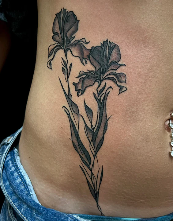 A delicate black and grey fine-line tattoo on a person's side abdomen featuring two elegant iris flowers. The design uses soft, translucent shading and flowing lines to create a graceful, minimalist botanical illustration next to a belly button piercing