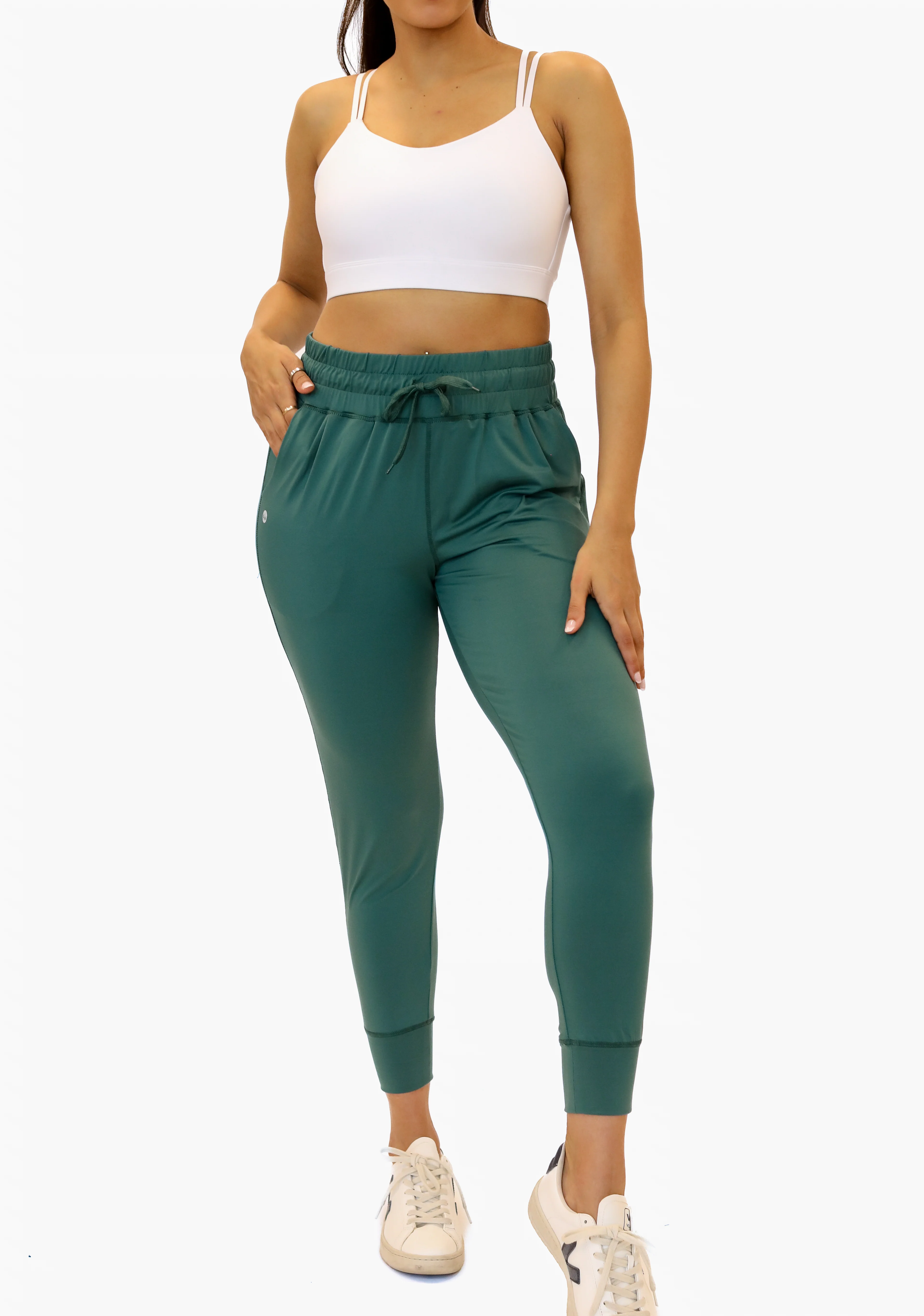 A close-up showing the fit of a model wearing Senita high-waisted, tapered joggers in a muted sage green color featuring a smocked drawstring waistband, deep side pockets, and fitted ankle cuffs, paired with a white sports bra and white sneakers.