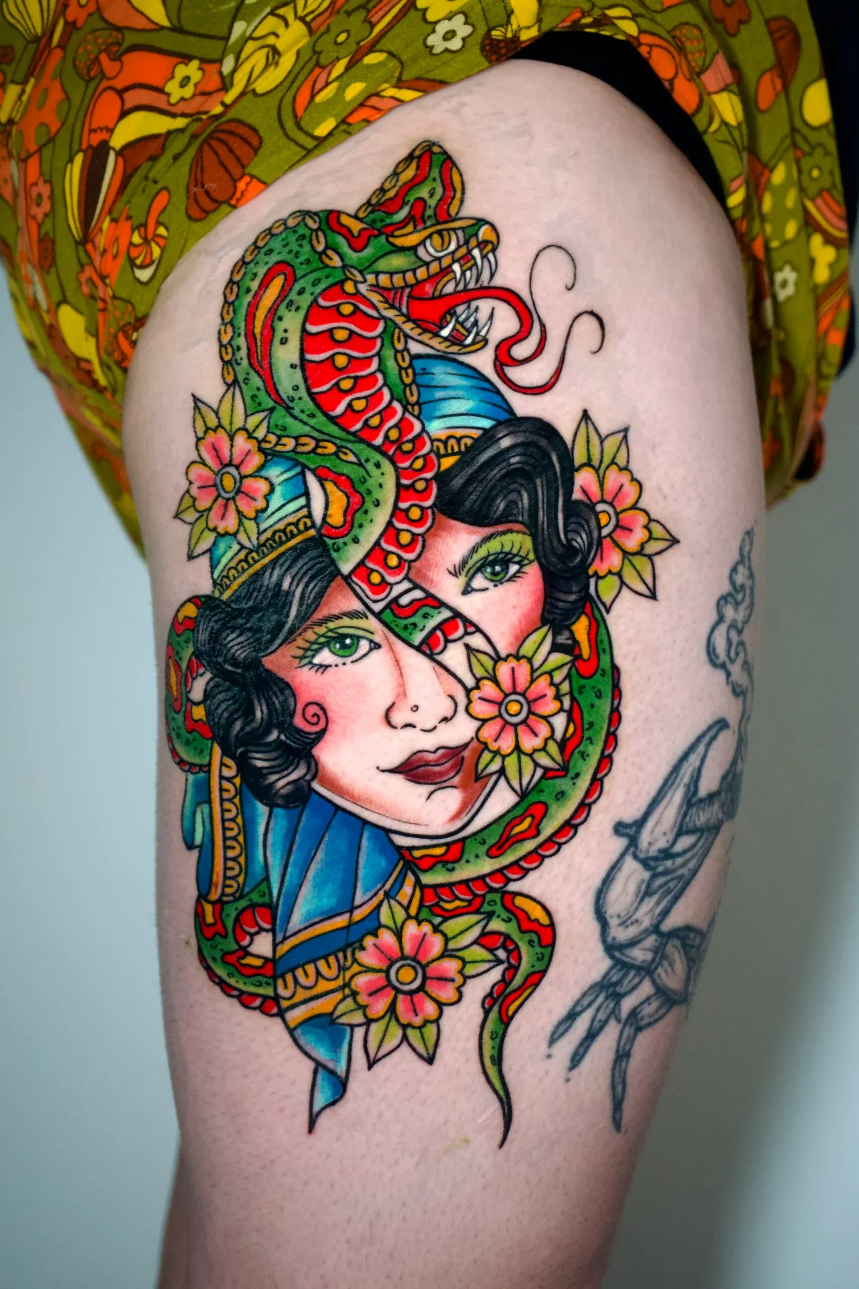 Vibrant traditional-style thigh tattoo of a woman's face wrapped in a colorful snake with flowers.