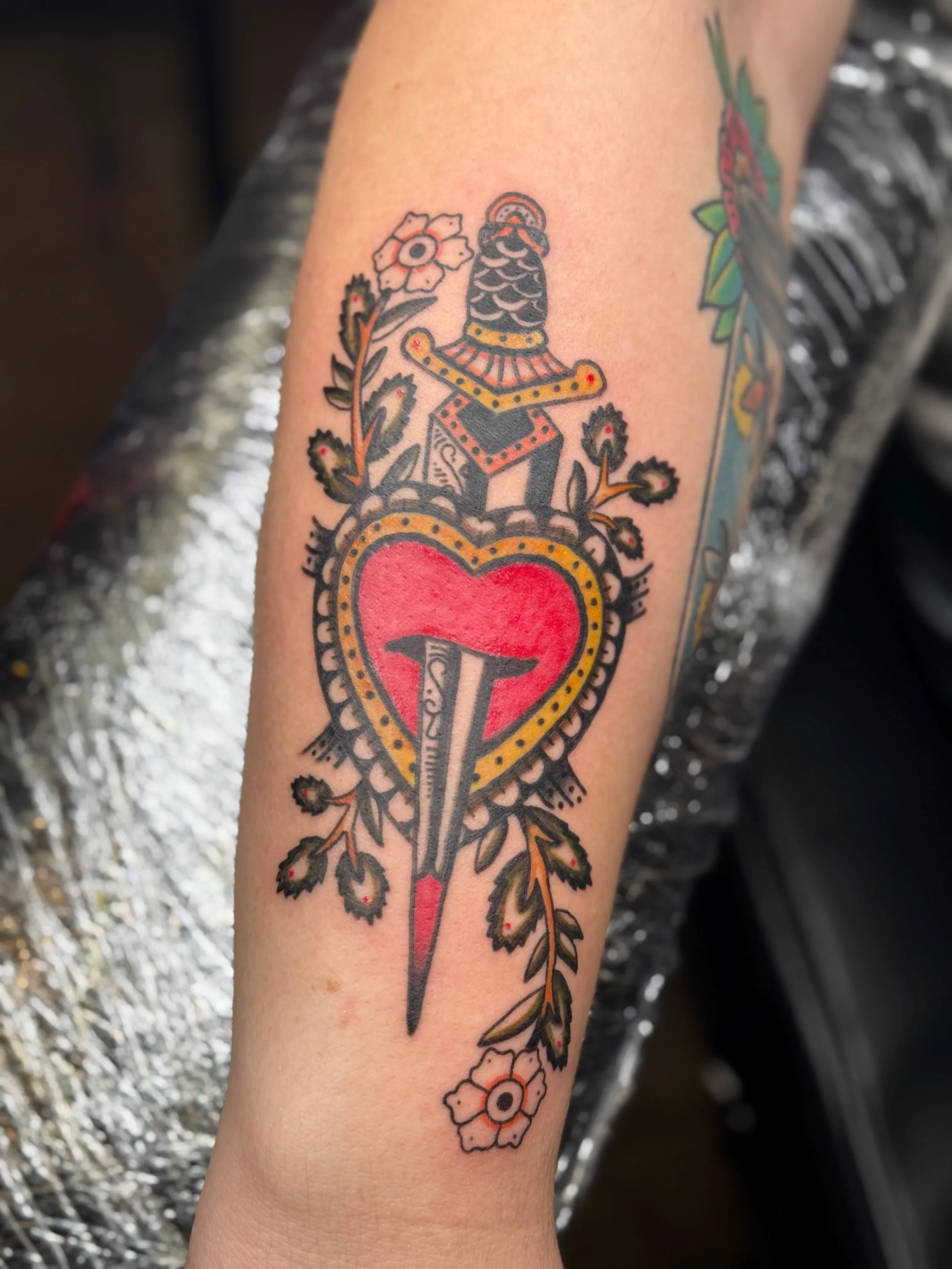 Traditional style tattoo of a dagger piercing a bright red heart surrounded by leaves and flowers.