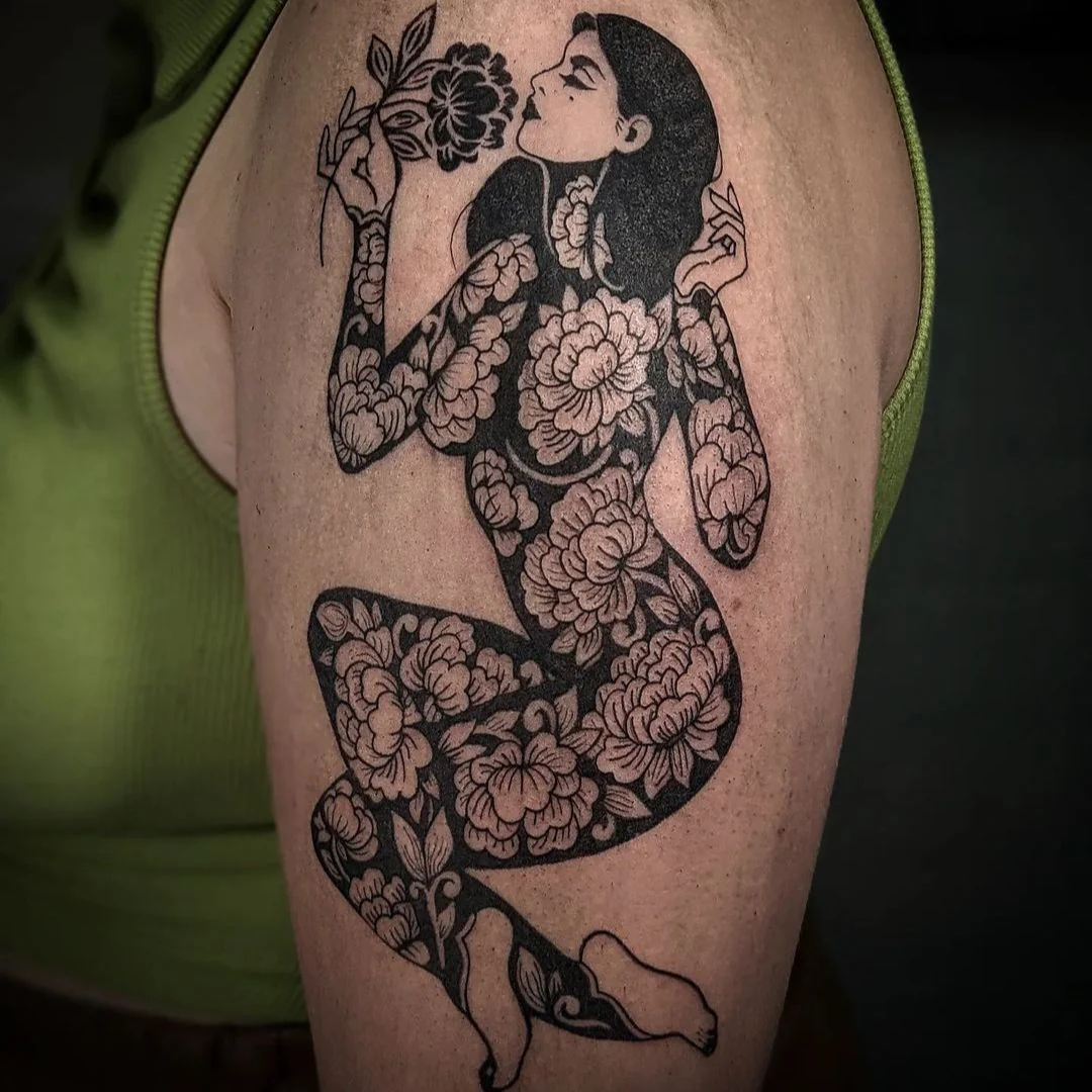 A blackwork tattoo on an upper arm depicting the silhouette of a kneeling woman whose entire body is filled with a detailed pattern of blooming peony flowers.