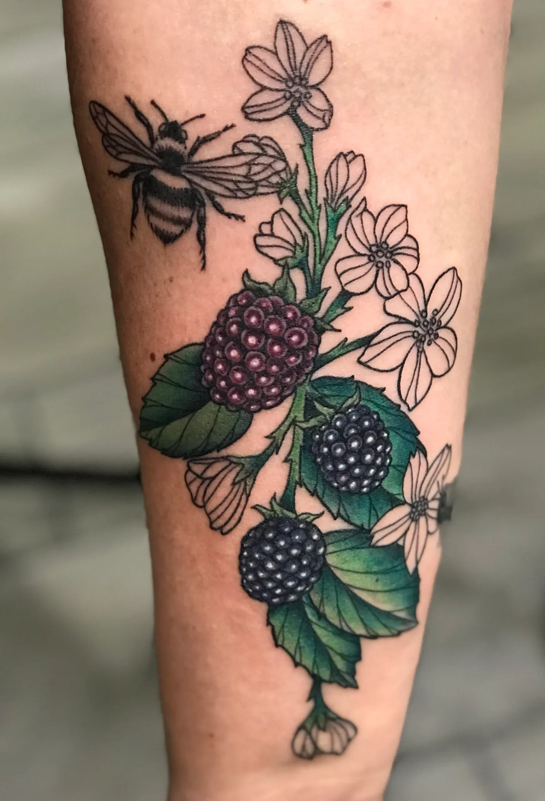 Detailed forearm tattoo of ripe blackberries, delicate white flowers, green leaves, and a bee.