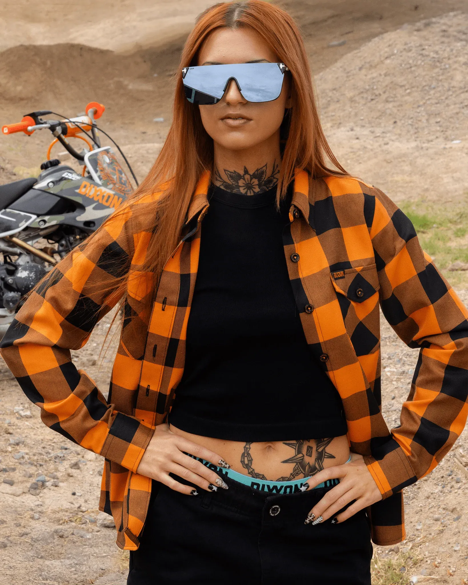 Tattooed woman in sunglasses wearing an unbuttoned orange and black Flat Track Dixxon flannel outdoors near a pit bike.