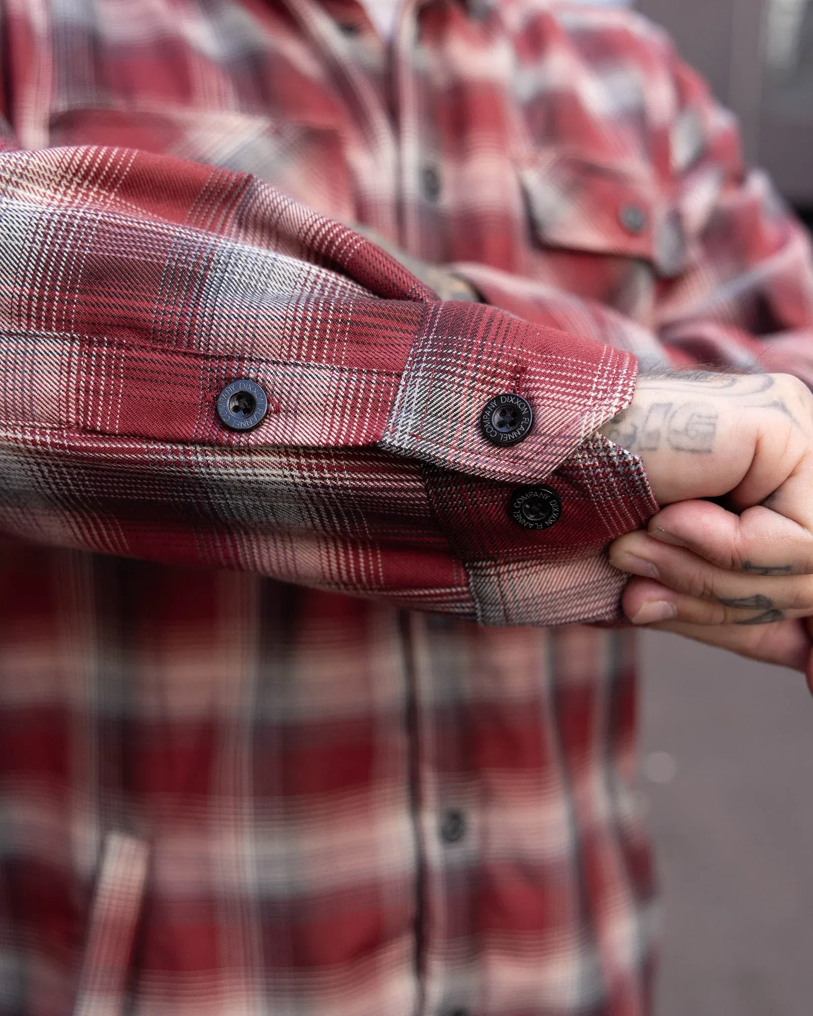 Close-up of a red and gray plaid flannel jacket sleeve showing custom Dixxon button cuffs and heavy stitching.