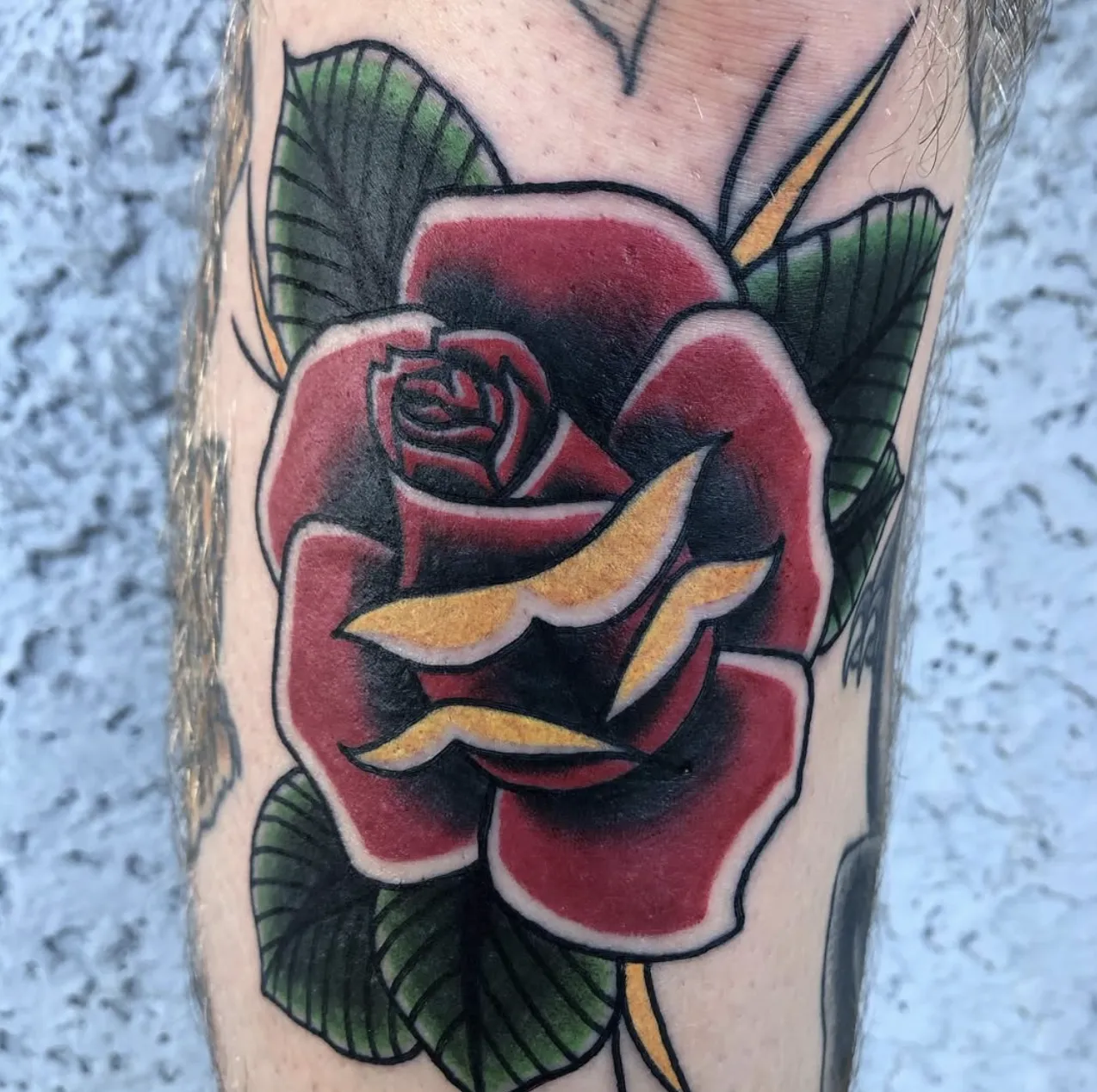 American Traditional red rose tattoo with bold black shading, yellow highlights, and green leaves.