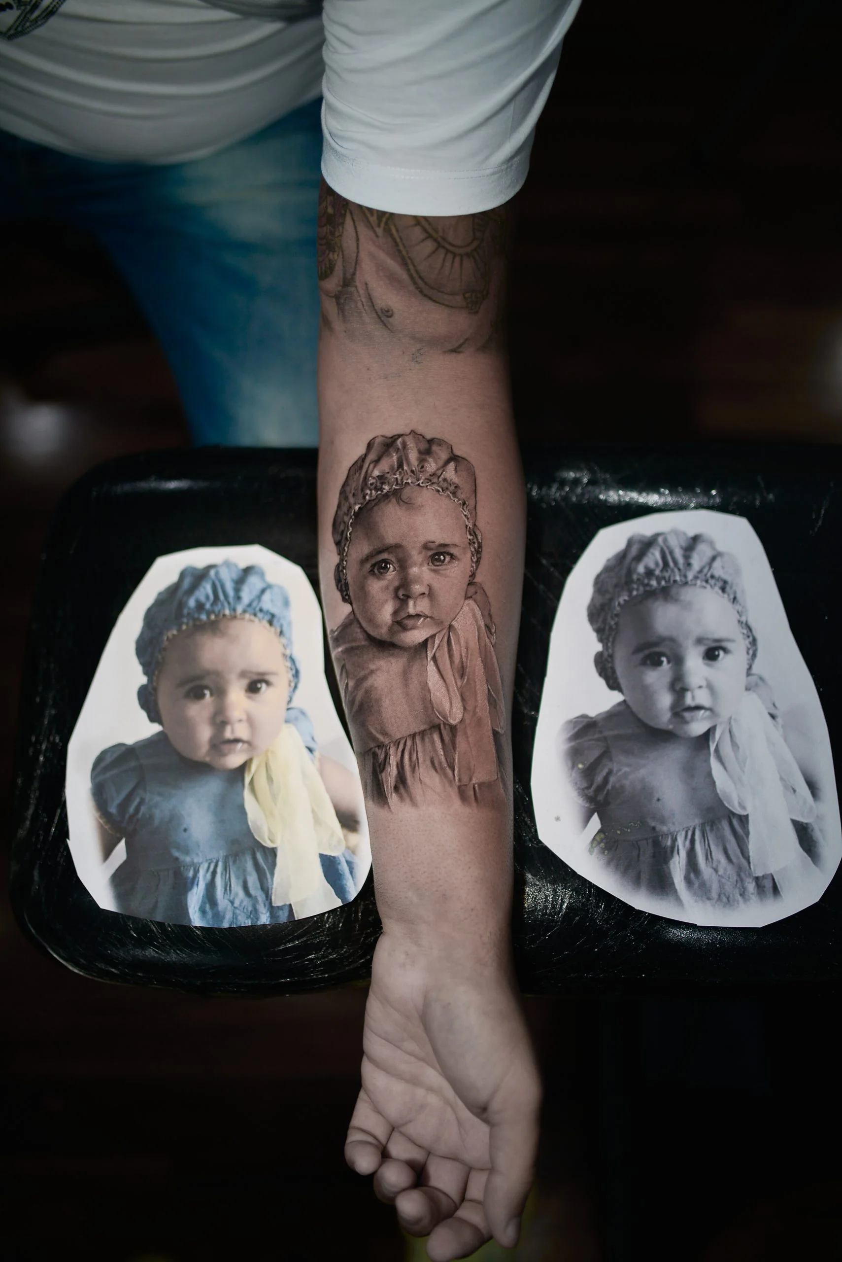 A hyper-realistic black-and-grey portrait tattoo of a baby wearing a bonnet, inked on a person's forearm. The arm is positioned between two printed reference photographs of the same baby used by the artist to achieve the photographic detail.