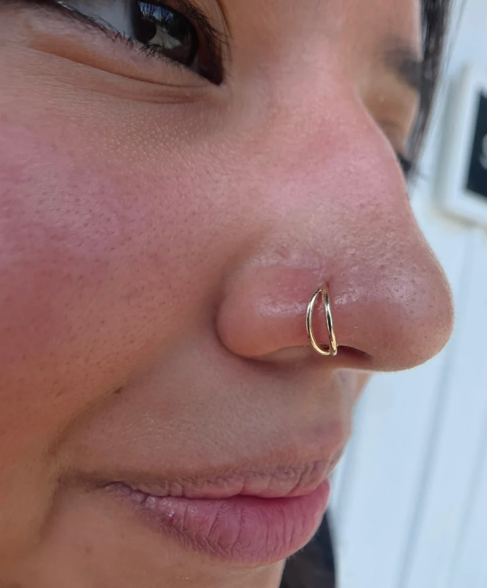 Side profile close-up of a client showing a double-layered gold hoop nostril piercing on the side of the nose.