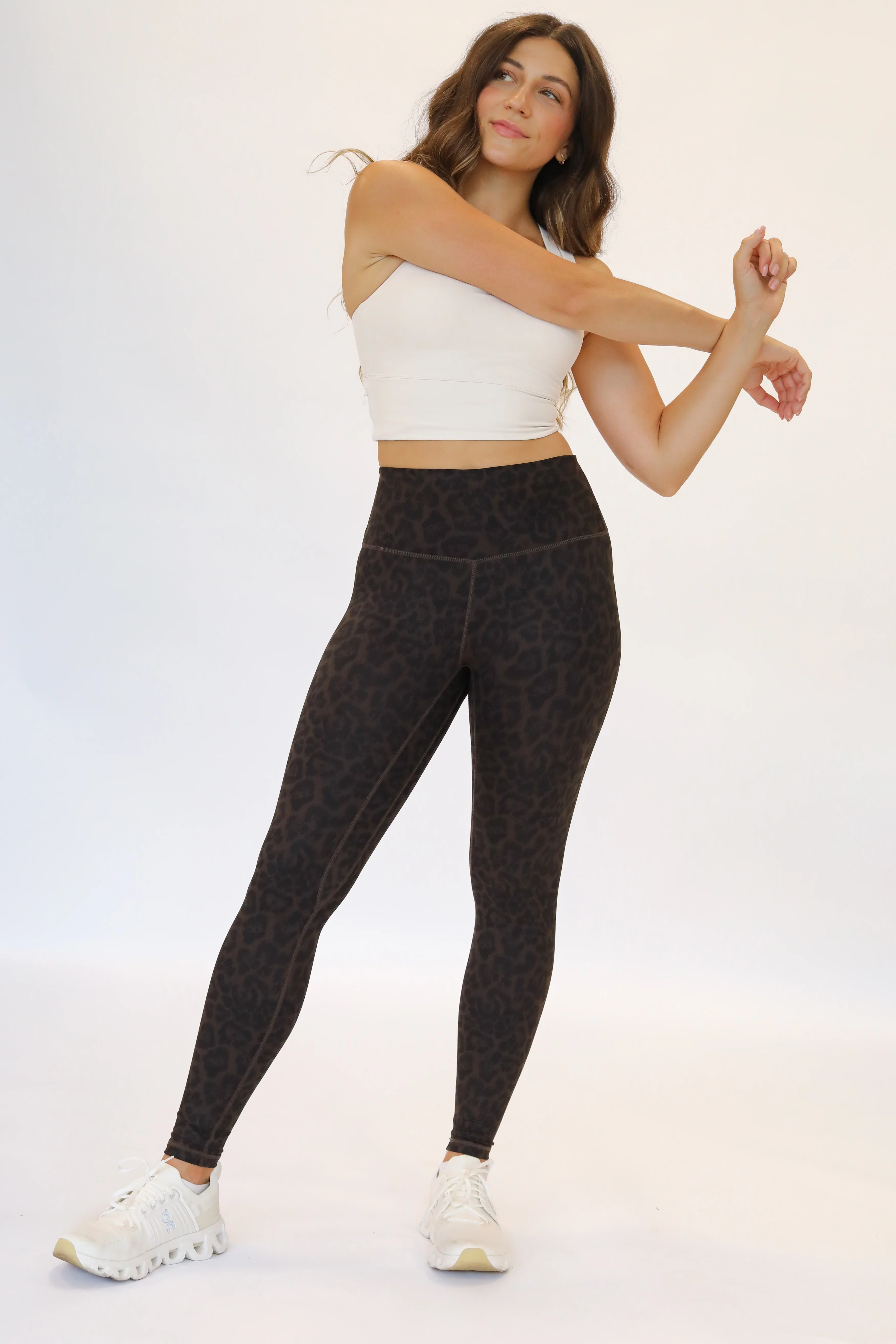 Full-body shot of a woman stretching her arms while wearing dark cocoa leopard print high-waisted leggings paired with a cream-colored cropped sports bra top and white athletic sneakers.