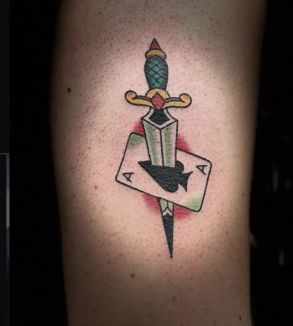 American Traditional dagger tattoo piercing through an Ace of Spades playing card with red background shading.