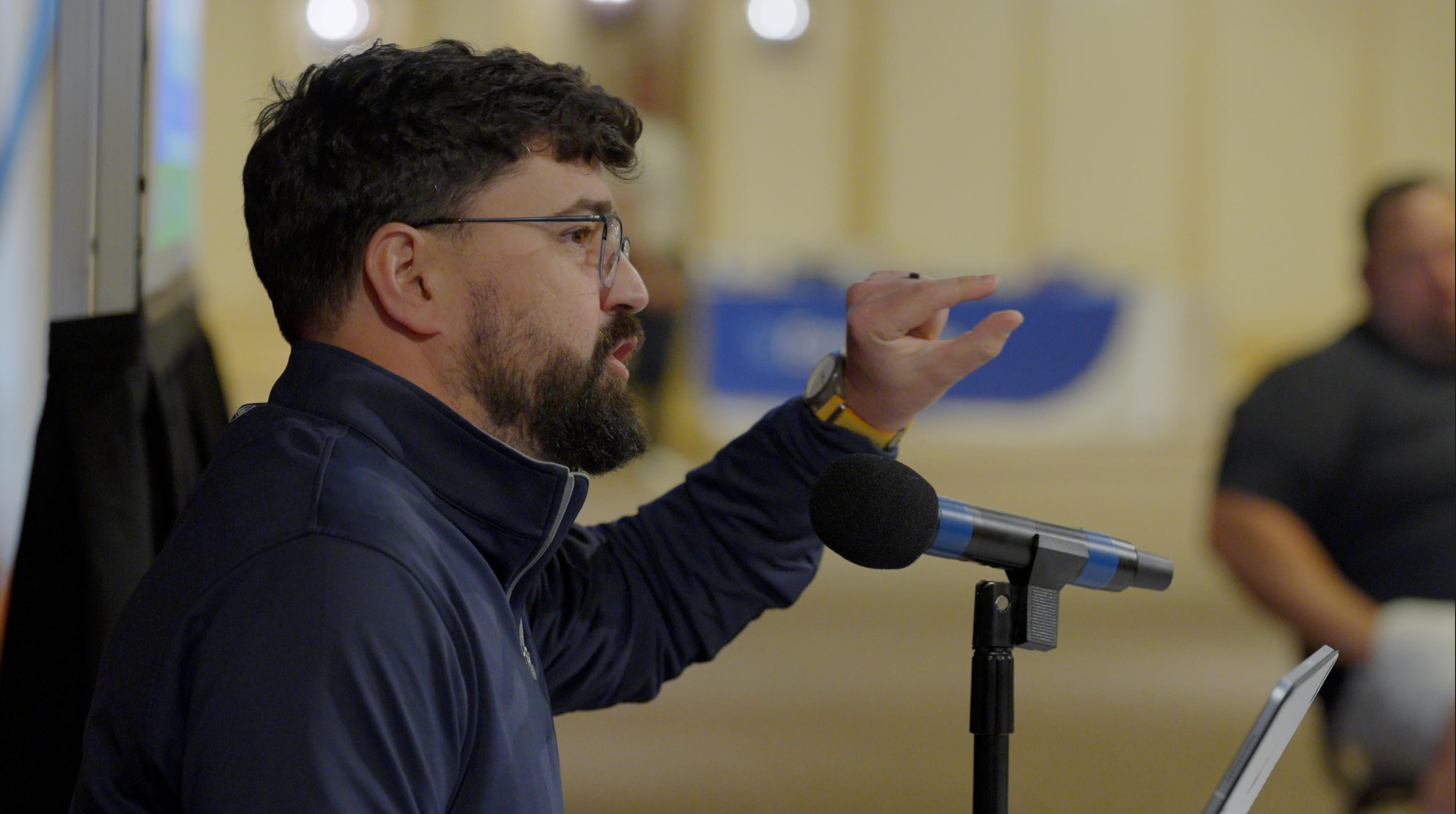 Man with glasses and beard gestures while speaking into a microphone