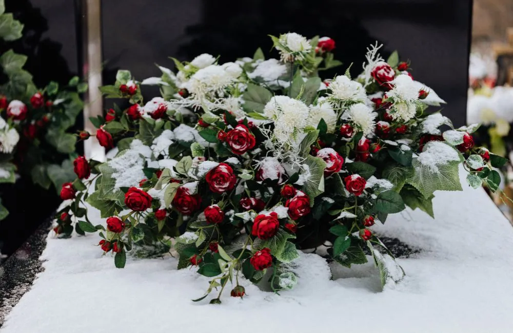 A floral arrangement featuring artificial or preserved red roses and white flowers with green foliage, dusted with a layer of snow, possibly created using a snow machine for a winter display or event.