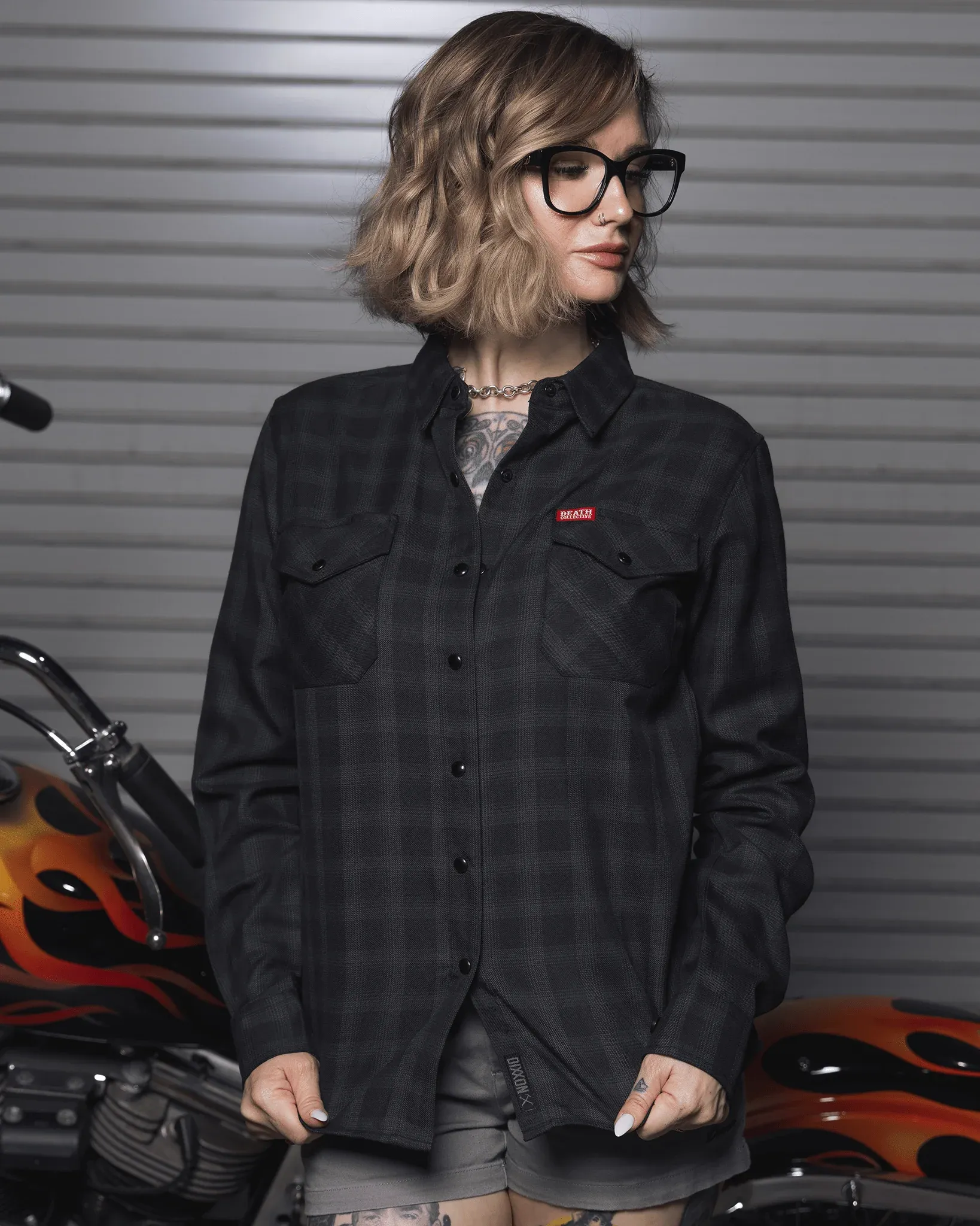Blonde woman wearing a dark gray Death Collective x Dixxon flannel in front of a custom flamed motorcycle.