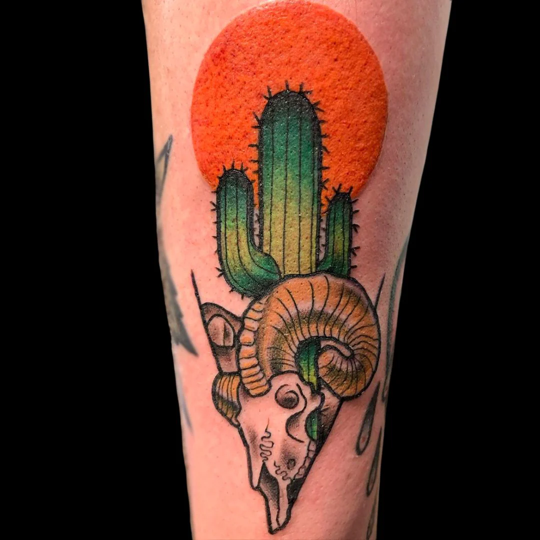 A vibrant color tattoo on a person's leg depicting a green saguaro cactus growing from a ram's skull. A large, solid orange sun sits in the background, creating a bold desert-themed composition.
