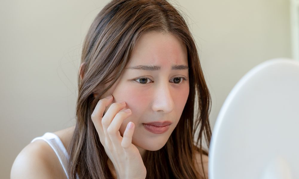 A concerned woman looking in a mirror to examine redness and irritation on her face, wondering if natural remedies like tallow for skin are better for skin barrier repair.