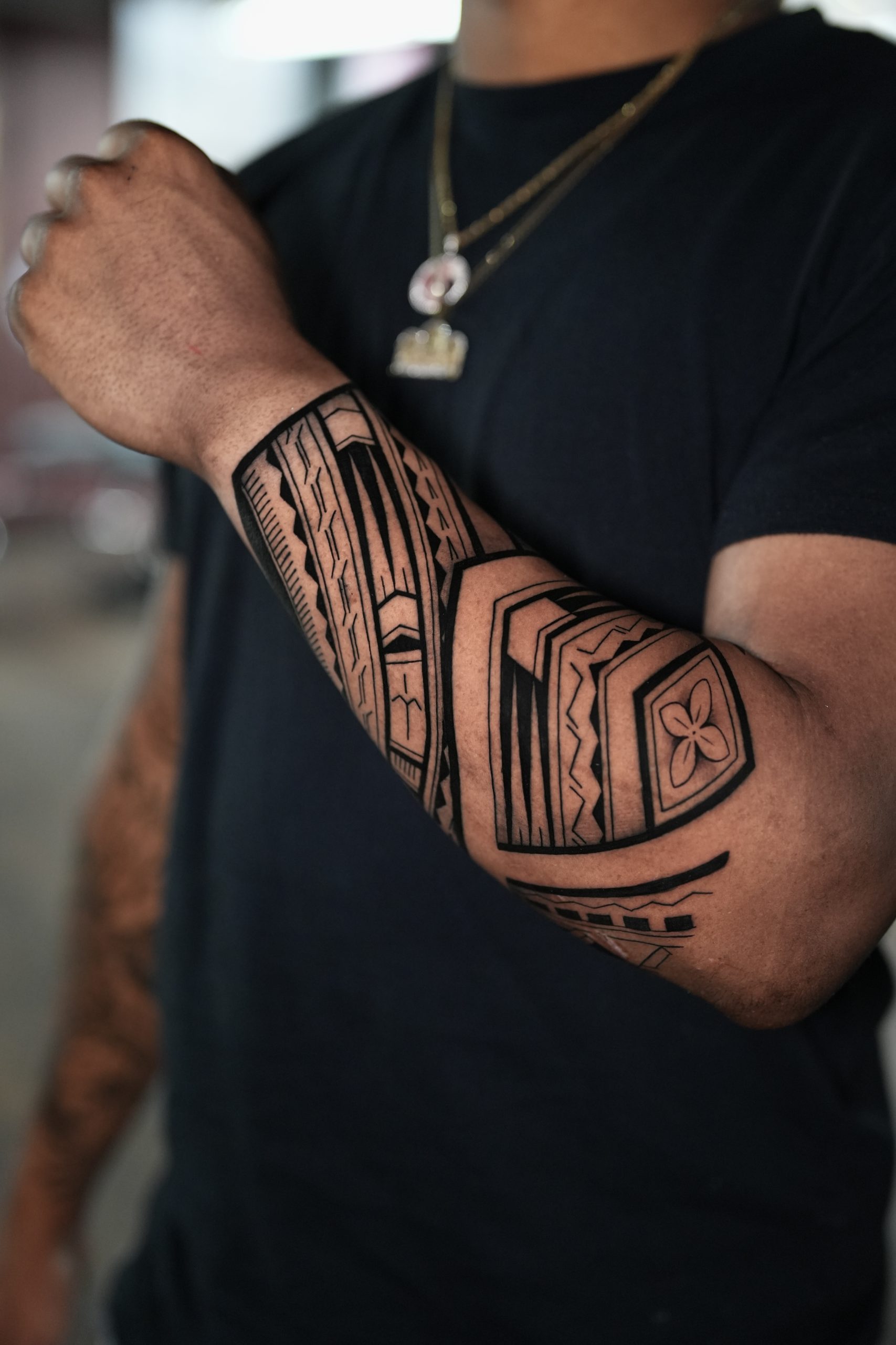 A bold, heavy blackwork Polynesian-inspired tribal tattoo sleeve wrapped around a man's forearm and inner elbow, featuring sharp geometric lines, chevrons, and a central four-petal floral design element.