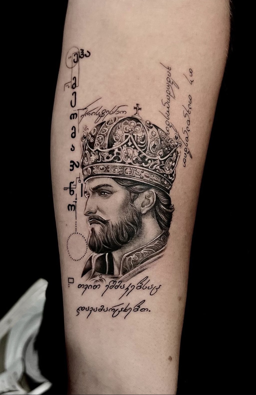 A precise black-and-grey micro-realism portrait tattoo of a bearded king wearing an ornate, detailed crown, accented by elegant script lettering and abstract geometric linework on an arm.