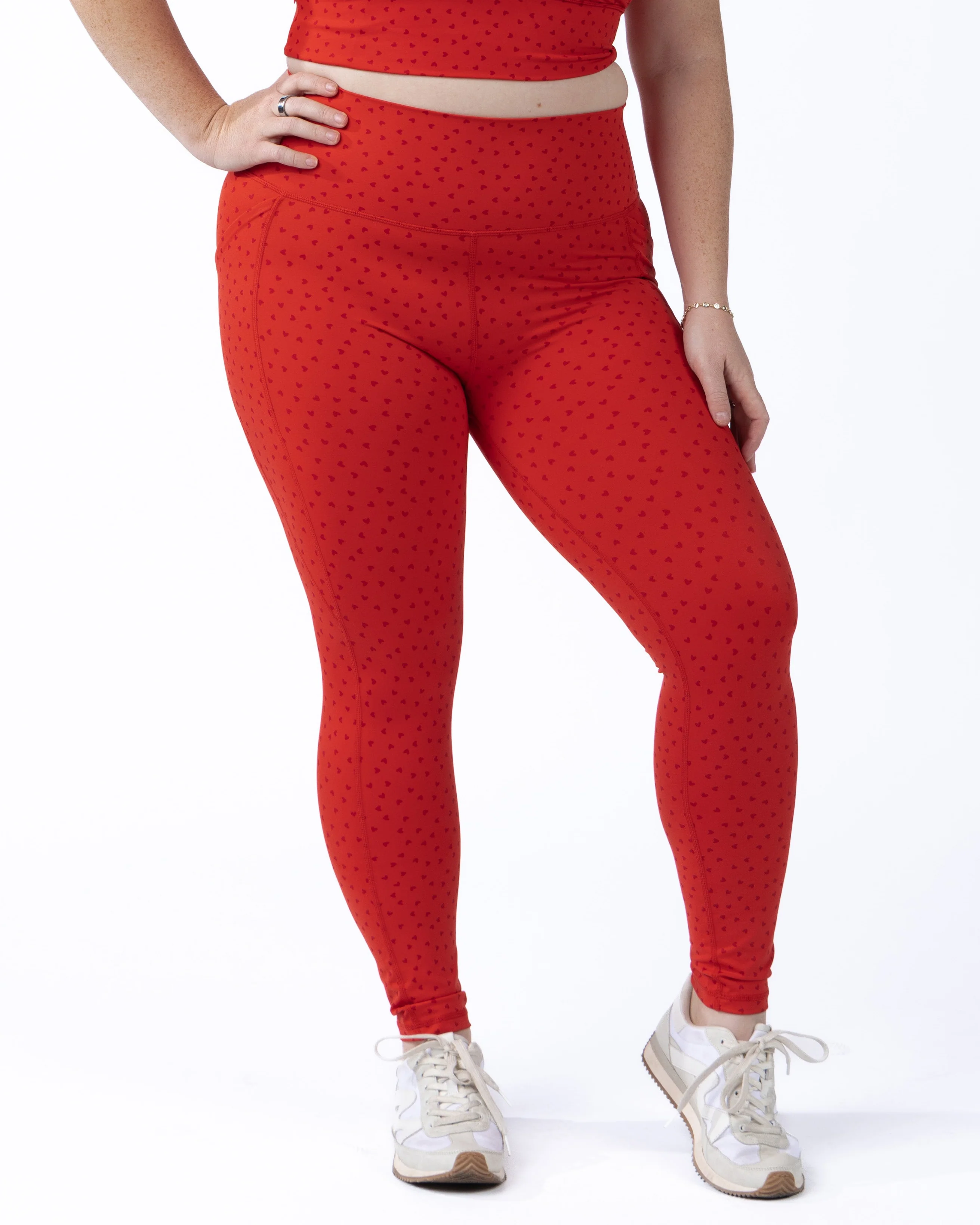 A detailed studio shot highlighting the inclusive fit of a model wearing Senita high-waisted performance leggings in bright red with a subtle, all-over mini heart pattern, complete with functional side pockets and styled with white sneakers.