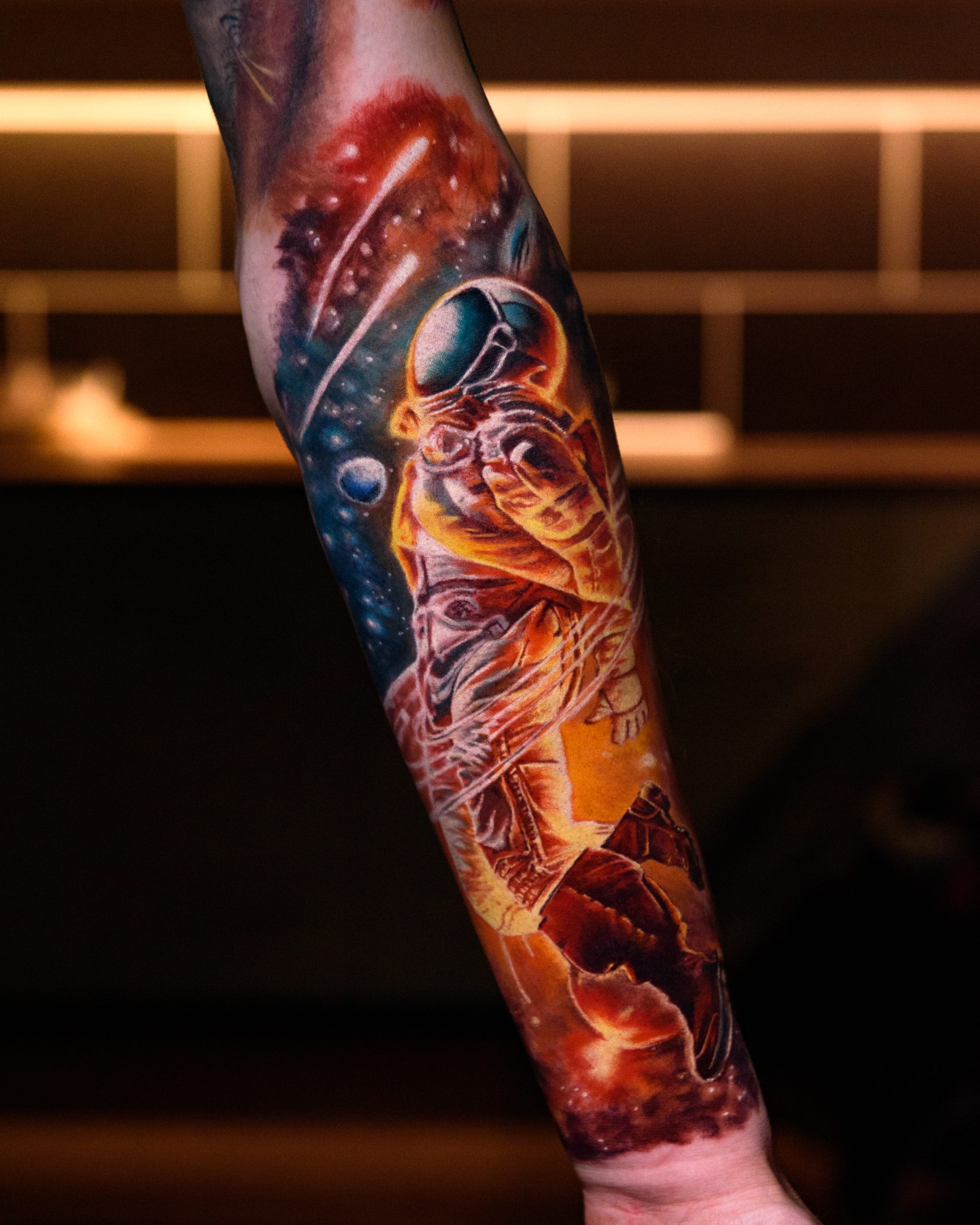 A vibrant, full-color realism tattoo sleeve on a forearm featuring an astronaut floating in deep space, surrounded by swirling cosmic nebulas in dramatic shades of orange, red, blue, and black.