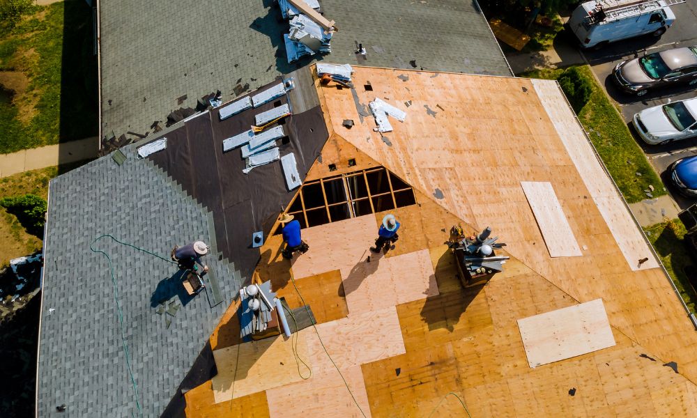 An aerial, top-down drone view of contractors performing a major roof replacement on a residential home, exposing the plywood deck and installing a brand new roof with fresh shingles and underlayment.