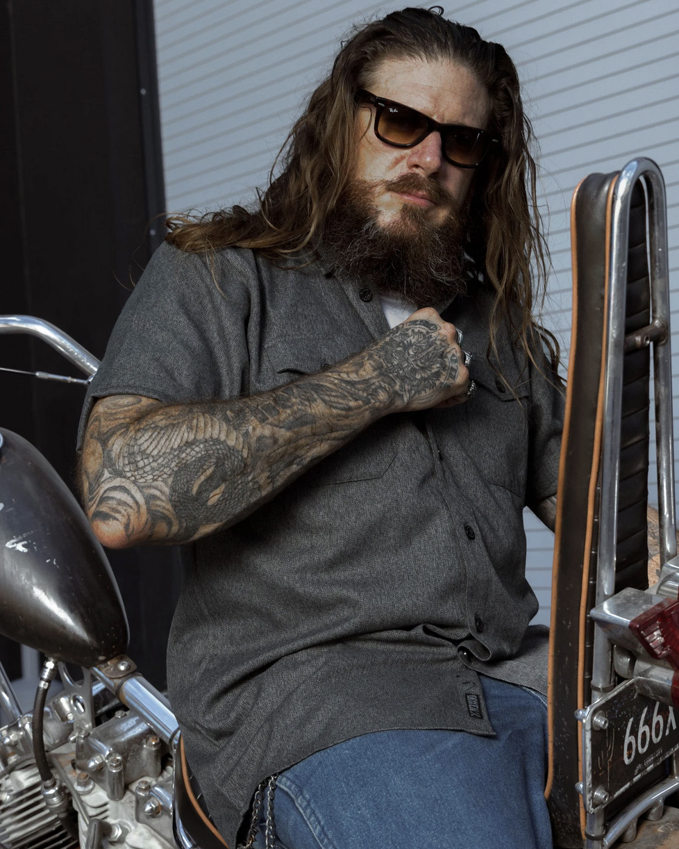 A man with long hair, a beard, and heavily tattooed arms sitting on a custom motorcycle. He is wearing a dark grey short-sleeve Dixxon work shirt, embodying the brand's deep roots in bike culture and absolute defiance.