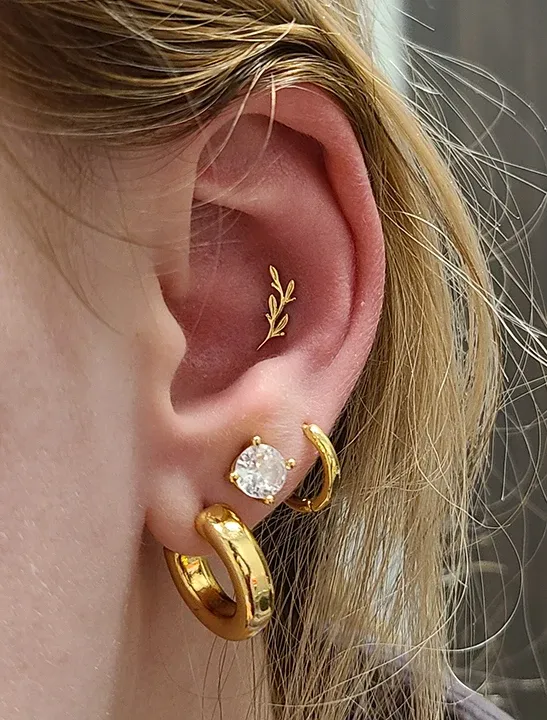 Ear curation with gold hoop earrings, a large crystal lobe stud, and a gold vine cartilage stud.