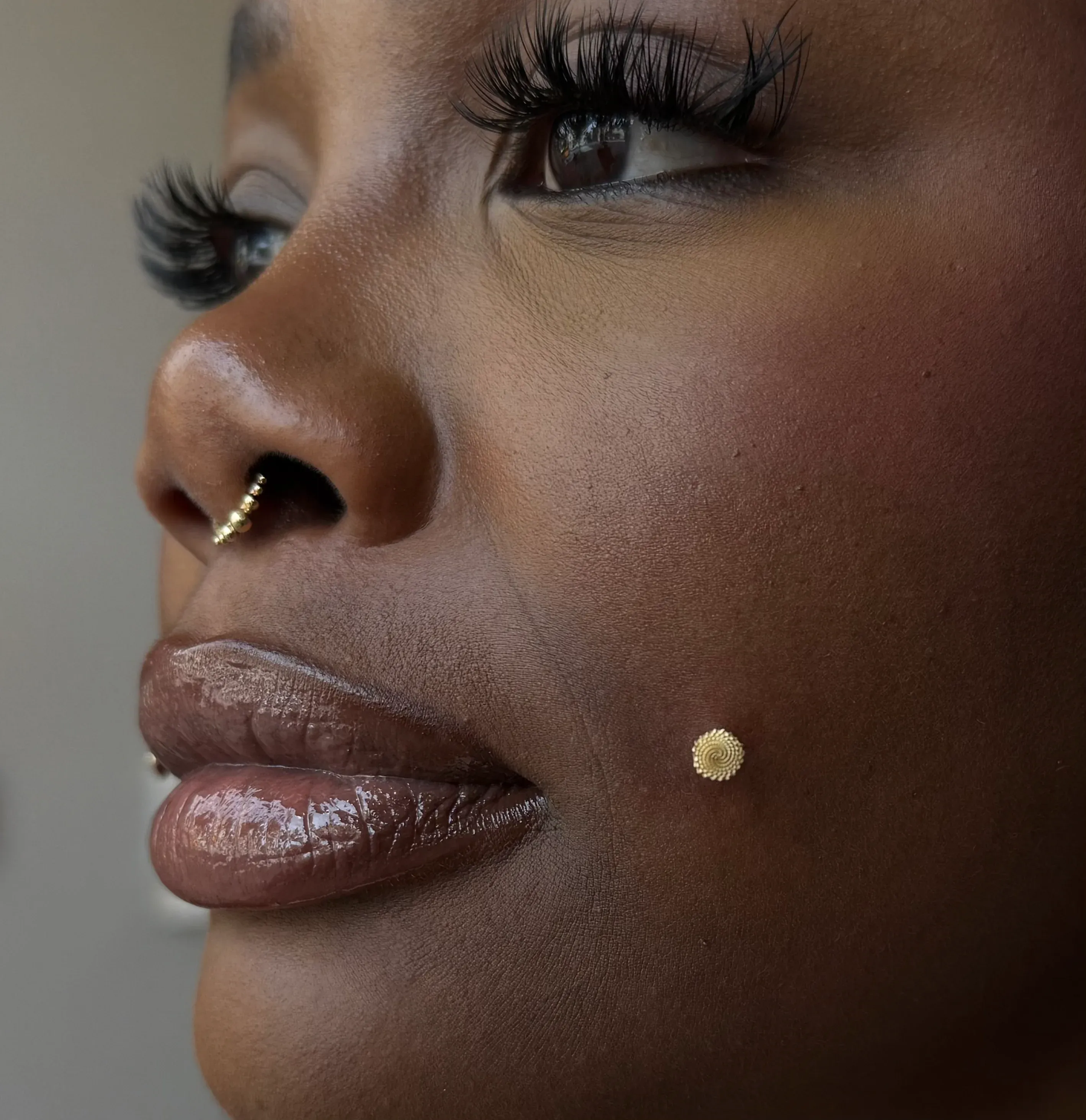 Close-up profile of a woman with dark skin featuring a gold beaded septum clicker and a textured gold cheek dermal piercing.
