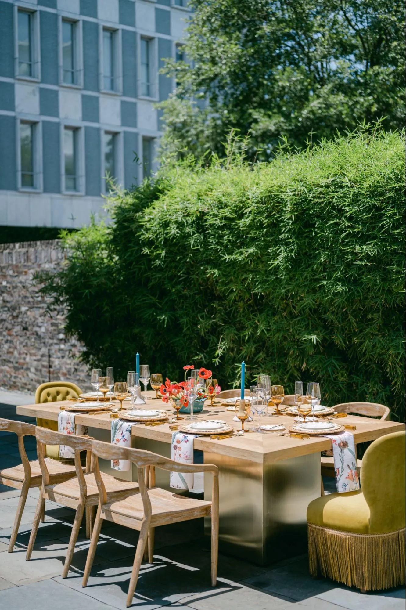 Outdoor birthday dinner setup with wood dining table, patterned runners, gold flatware, and upholstered accent chairs.
