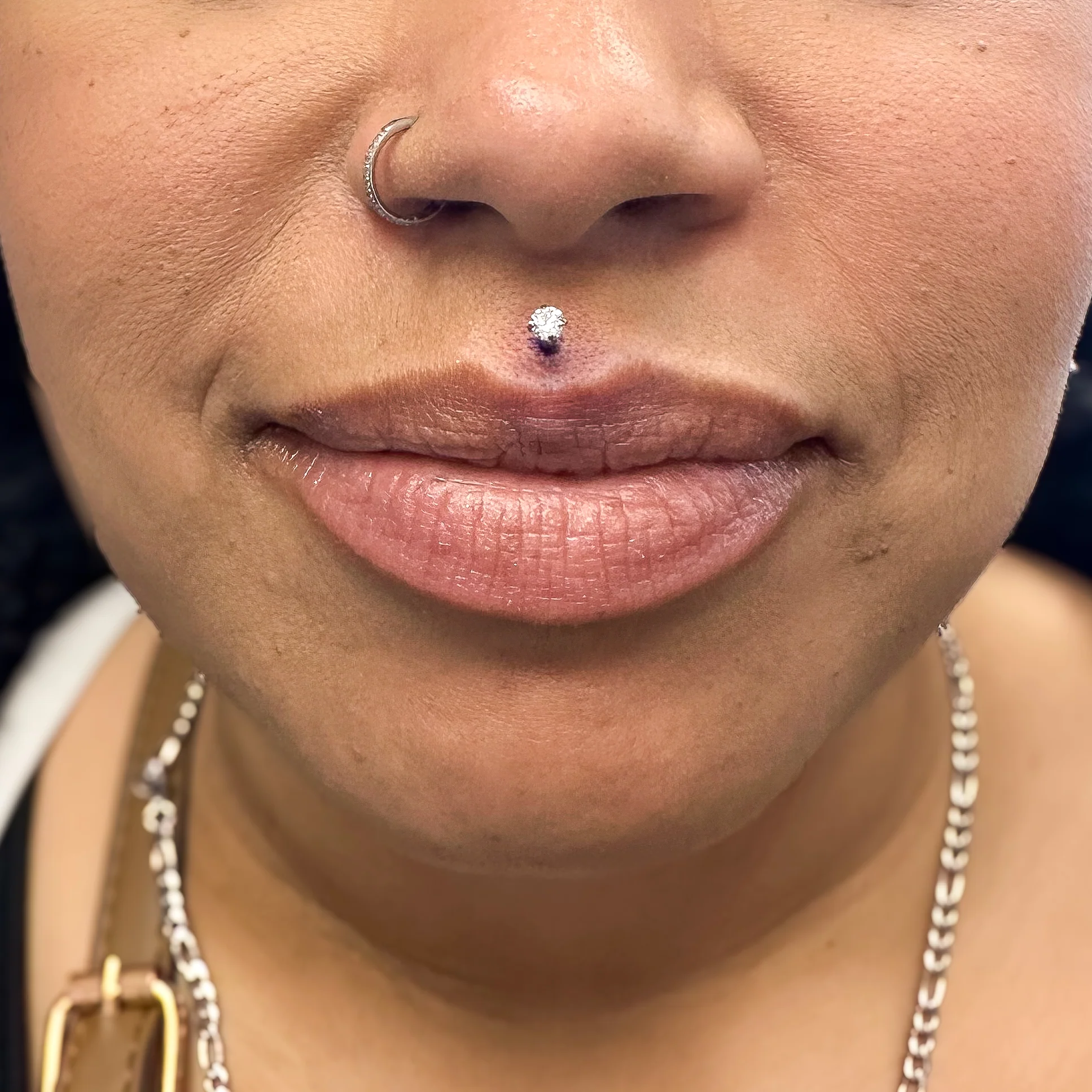 Close-up of a client showing a fresh philtrum piercing adorned with a sparkling diamond stud on the upper lip, alongside a seamless silver nostril hoop piercing.
