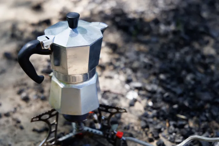 A silver Moka pot sits on a camping stove, ready to brew a hot cup of coffee in the wilderness.