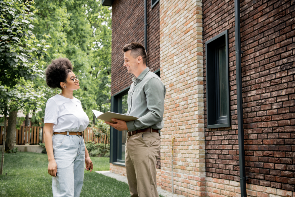 A professional inspector holds a folder while discussing a home inspection cost with a client in the backyard of a modern brick house.