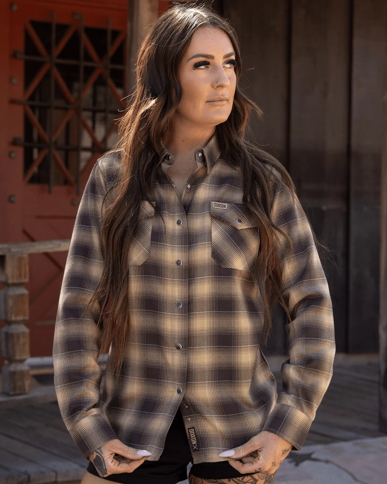 Woman wearing a tan and dark brown plaid Dixxon flannel with long dark hair and tattooed hands.
