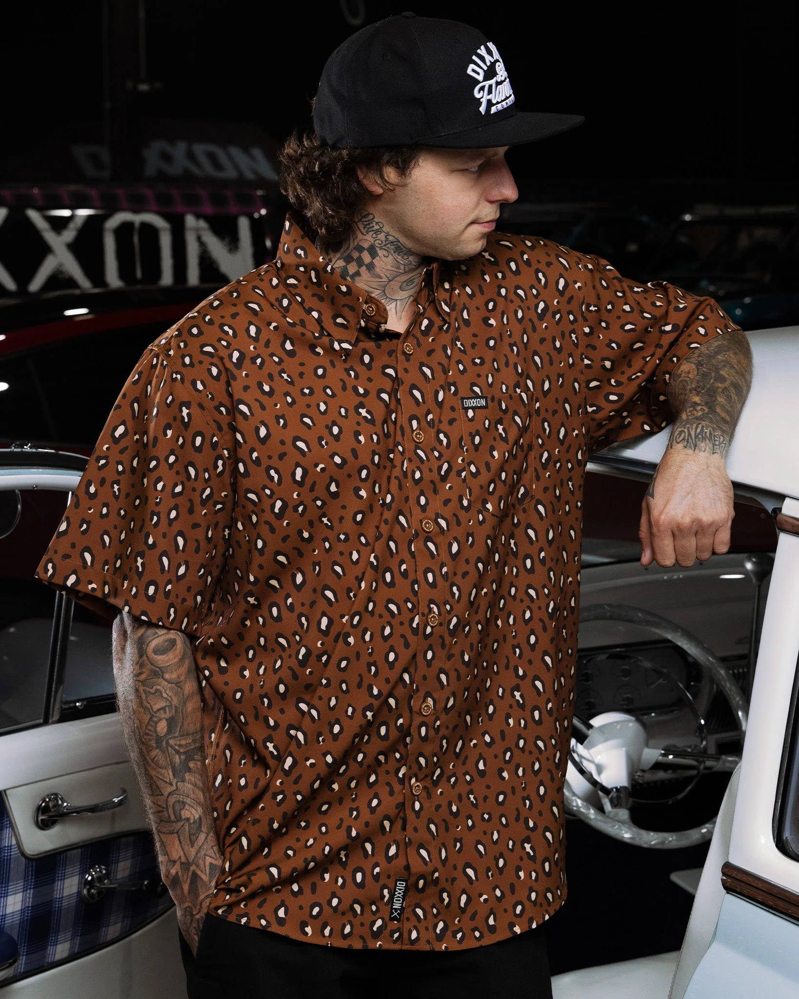 A tattooed man wearing a black flat-bill snapback hat leans against a vintage white vehicle, sporting a short-sleeve button-up shirt. The shirt features an all-over brown leopard or animal spot print pattern with a small black woven Dixxon tag visible on the chest pocket line.