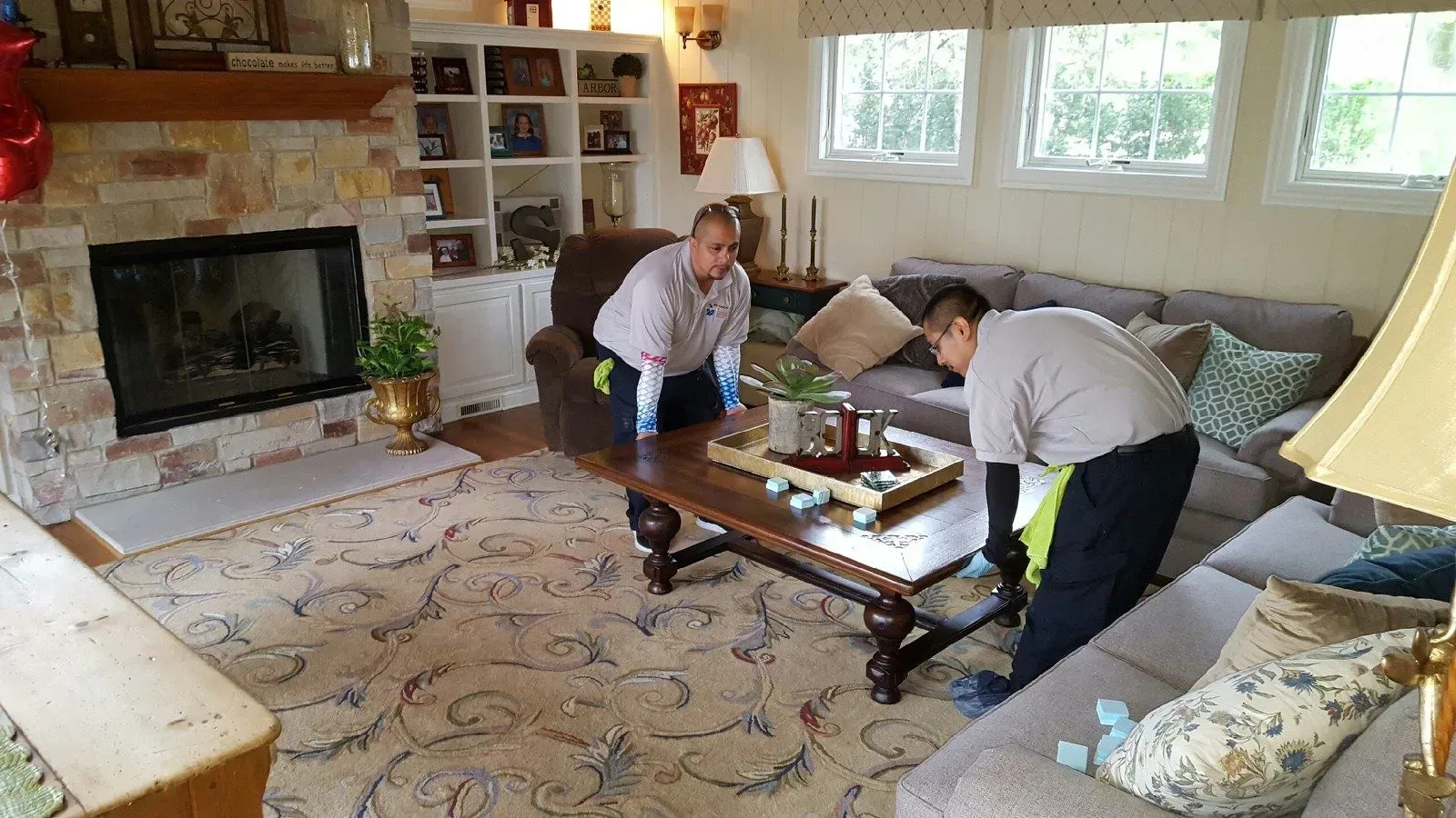 Two professional cleaners in uniforms lifting a wooden coffee table in a living room, preparing for upholstery cleaning, with a cozy fireplace and a well-decorated space in the background.