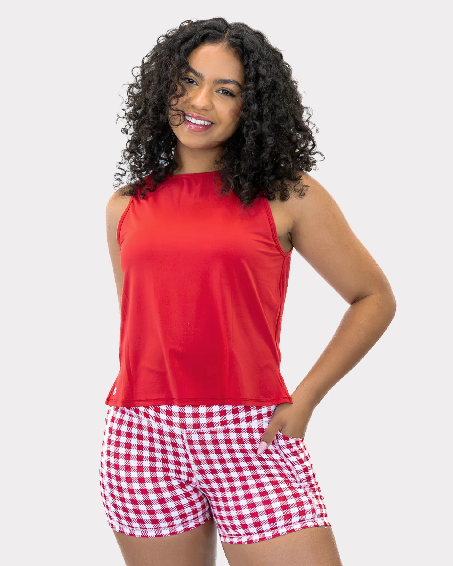 A smiling woman with dark, curly hair wears a solid red sleeveless crewneck top paired with high-waisted red and white gingham checkered shorts featuring side pockets.