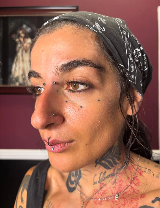 Side profile of a woman showcasing multiple facial piercings including a bridge piercing, nose stud, vertical labret lip ring, and decorative face tattoos