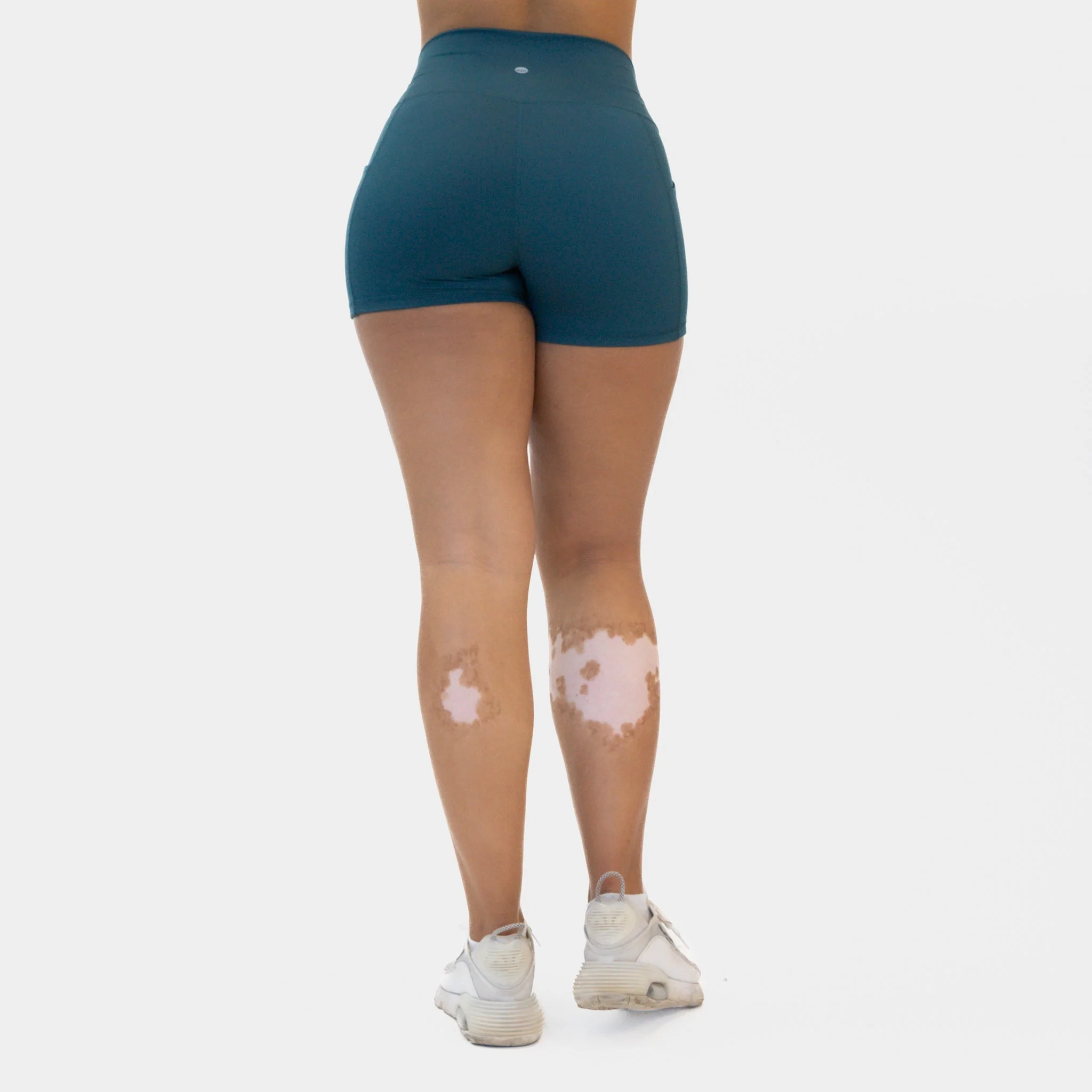 Rear view of a model from the waist down wearing teal, high-waisted compressive athletic shorts and white sneakers against a clean white background.
