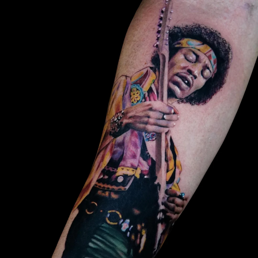 A highly detailed, realistic color portrait tattoo of Jimi Hendrix playing a guitar. The portrait captures his expression with closed eyes, wearing his iconic patterned headband and a vibrant yellow and purple outfit against a solid black background.