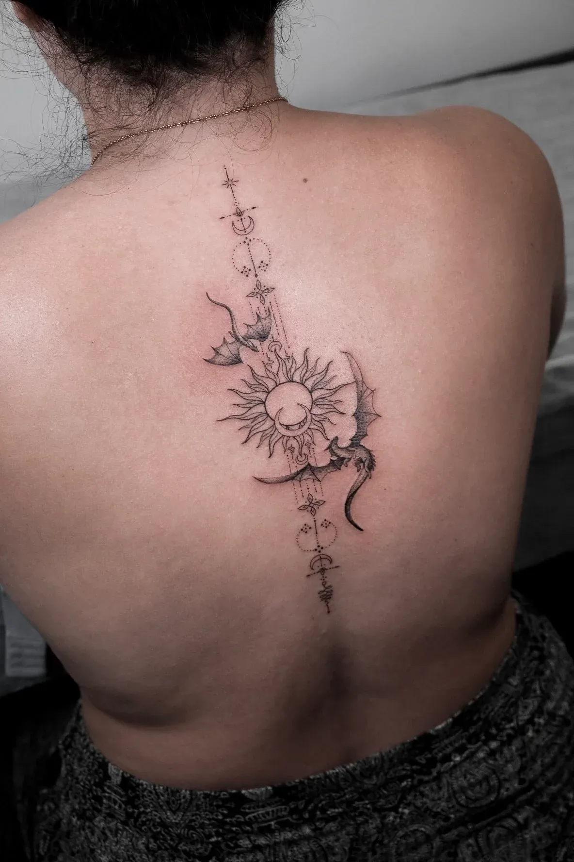 Fine-line back tattoo with celestial symbols, a sun, crescent moon, and flying dragons.