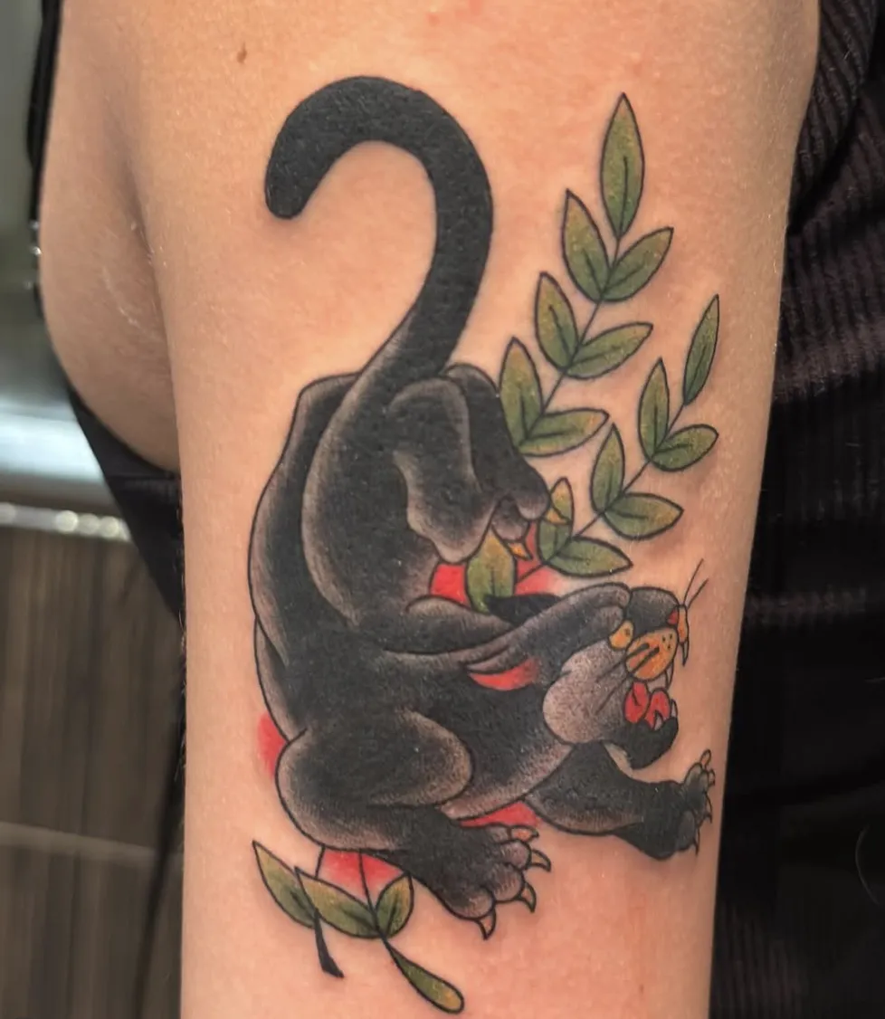 American Traditional black panther tattoo crawling downward beside green leafy branches and red background shading.
