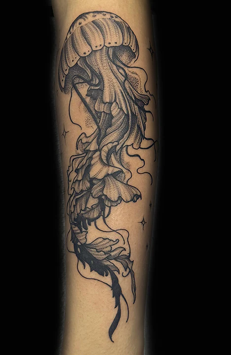 A detailed black and grey illustrative tattoo on a person's forearm featuring a large jellyfish. The design showcases intricate dot work on the bell and long, flowing tentacles intertwined with delicate star-like sparkles.