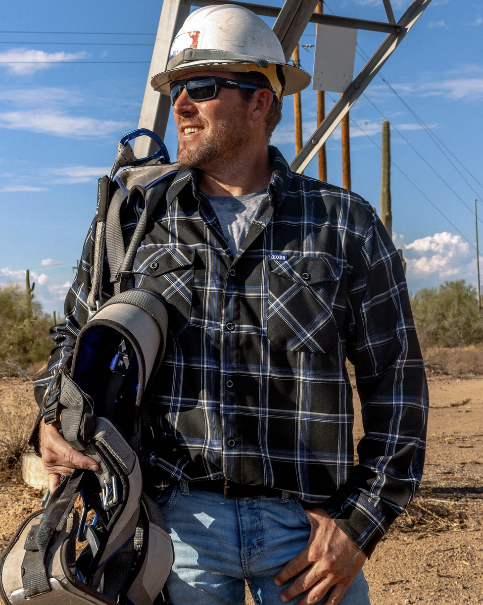 A smiling worker in a white hard hat and sunglasses wearing a blue, white, and black Dixxon flannel. He is carrying heavy-duty climbing harnesses in a desert landscape near a power line tower, illustrating the product's durability on the job site.
