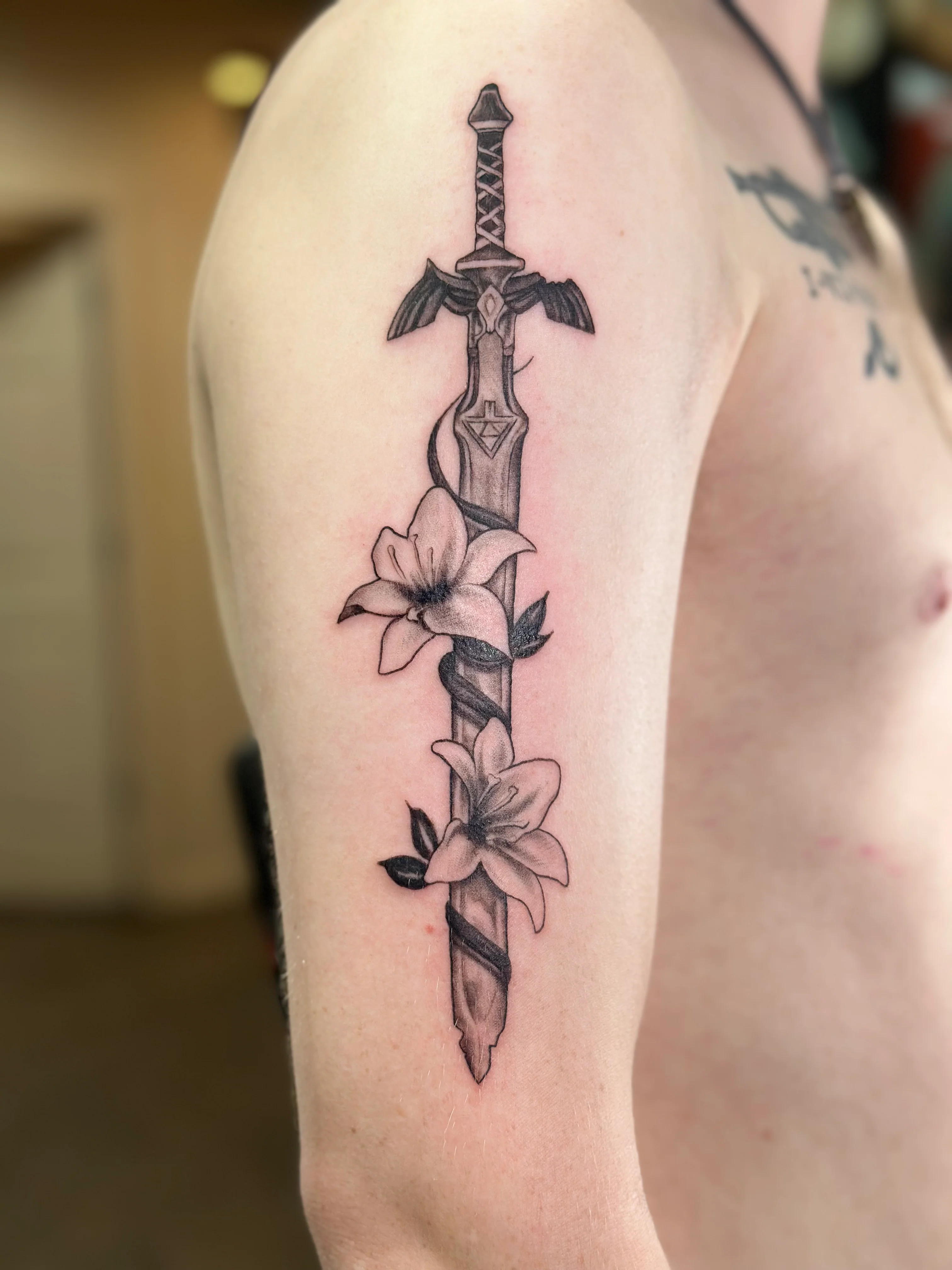 A black and grey illustrative tattoo on a person's upper arm and shoulder depicting a stylized fantasy sword resembling the Master Sword. Two detailed white lily flowers with dark green leaves are elegantly wrapped around the blade