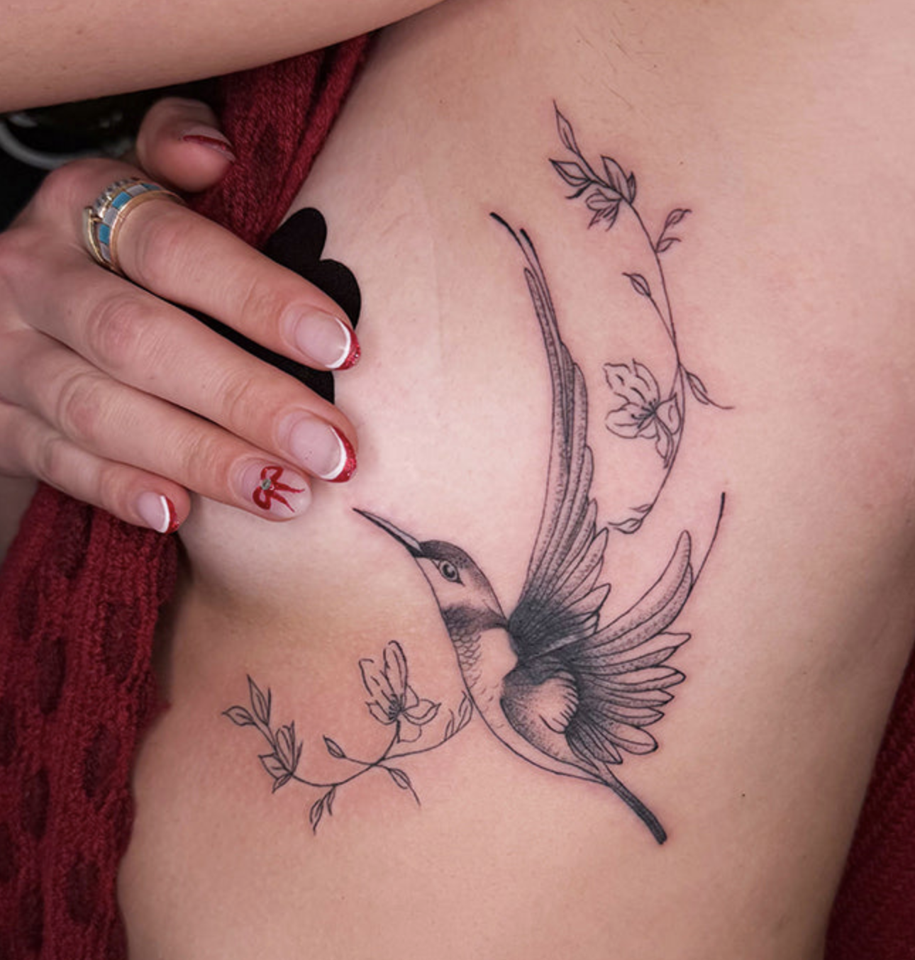 A detailed, black and grey fine-line tattoo on a woman's ribs, just under the bust. It depicts a hummingbird in mid-flight surrounded by delicate, flowing vines of small flowers and leaves.