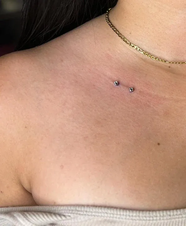 Collarbone dermal anchor piercing with two silver studs next to a gold chain necklace.