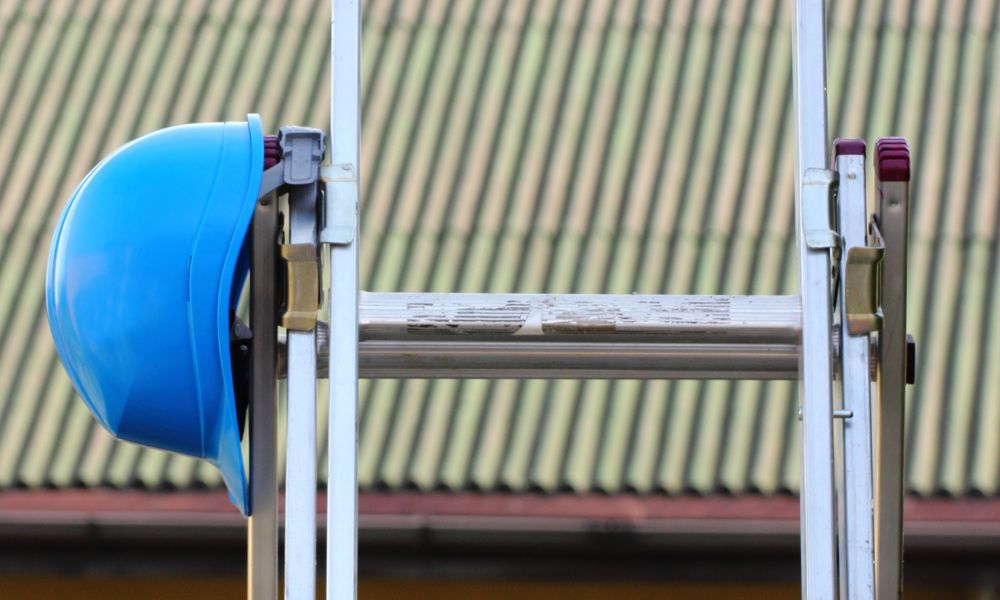 A blue safety hard hat hanging on an aluminum extension ladder during a residential roofing and maintenance project, with a roof visible in the blurred background.